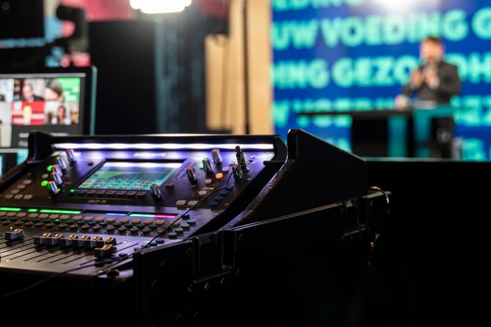 Digital mixing console at a live event, with a speaker on stage out of focus behind it