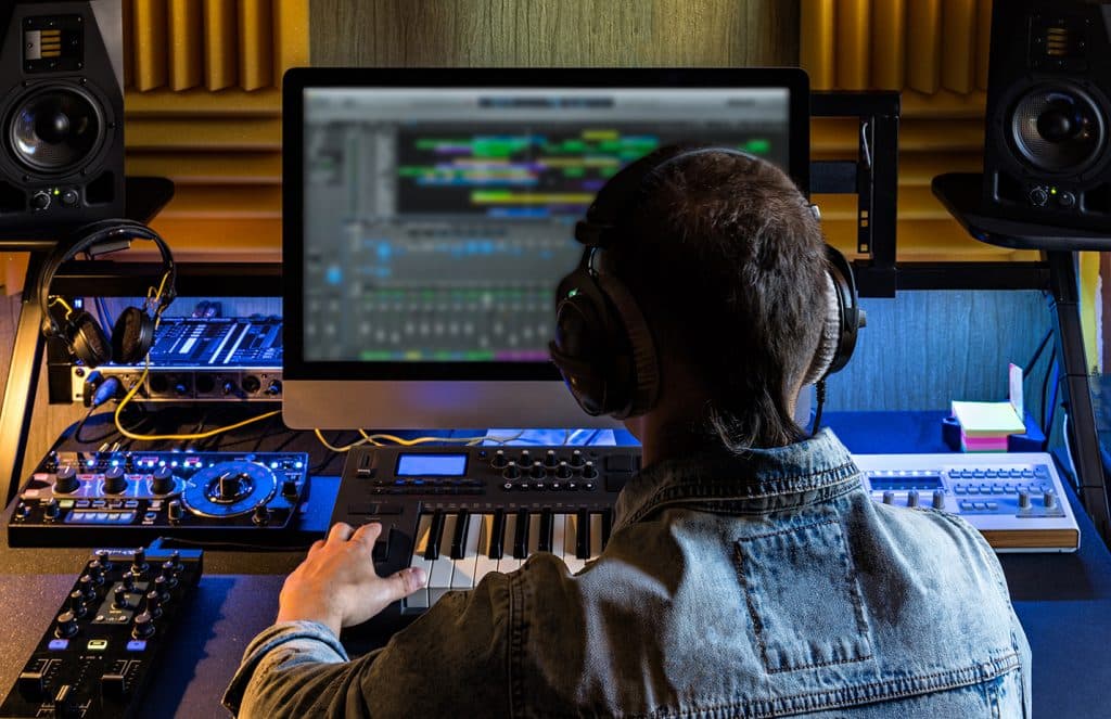 Producer wearing headphones playing a keyboard in front of a recording session on screen